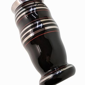 May include: A black vase with silver and red stripes. The vase has a glossy finish and a flared base. The top and bottom rims are silver. The stripes are horizontal and evenly spaced around the body of the vase.