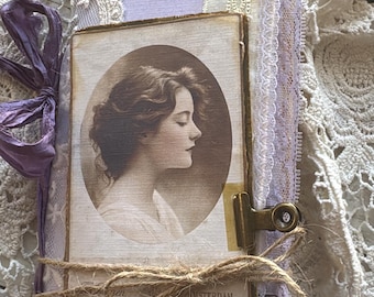 May include: A handmade journal with a light purple cover, decorated with layers of white lace and fabric trim. A vintage-style oval portrait of a person in profile is attached to the front with a brass clip and tied with a piece of brown twine.