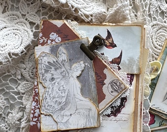 May include: A layered paper booklet featuring a fairy illustration, vintage-style script, and bird imagery, secured with a small metal clip. The booklet is placed atop a white crocheted lace fabric with intricate floral patterns and scalloped edges.