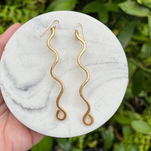 May include: A pair of gold-toned earrings with a wavy, abstract design. The earrings are displayed on a white marble surface.