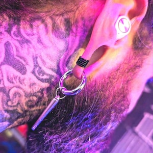 May include: A silver hoop earring with a dangling, cylindrical charm is shown. A black ring is on the ear. The ear has a white earbud. The background is a mix of purple and pink hues, with a tattoo visible.