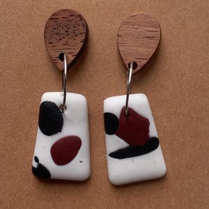 Puede incluir: Un par de pendientes colgantes. Cada pendiente presenta una parte superior de madera en forma de lágrima y una parte inferior rectangular blanca. La parte inferior blanca tiene formas abstractas negras y granates. Los pendientes cuelgan de aros plateados.