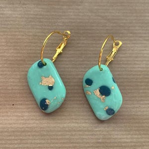 May include: A pair of gold hoop earrings with a light blue, rectangular pendant. The pendant has a speckled pattern of gold and dark blue.