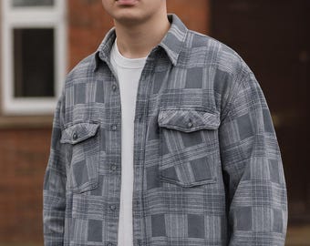 May include: A gray plaid button-down shirt with two chest pockets, worn over a white t-shirt. The shirt has a classic collar and long sleeves. The person is standing in front of a brick building.