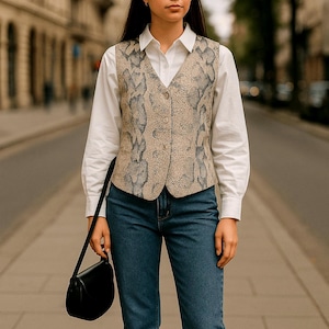 May include: A woman in a snakeskin print waistcoat, white shirt, and blue jeans, carrying a black shoulder bag. The outfit is suitable for various occasions. The woman is standing on a paved street.