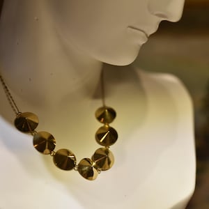 Puede incluir: Un collar dorado con una serie de colgantes circulares facetados. El collar se exhibe en un maniquí blanco, destacando su diseño y textura. Los colgantes están dispuestos en un patrón graduado, creando una pieza llamativa.