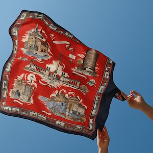 May include: A red silk scarf with a navy blue border, featuring illustrations of landmarks. The scarf has a repeating pattern of buildings and monuments, with the word "Budapest" on it. The scarf is being held up against a blue sky.