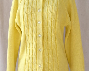 May include: A pale yellow cardigan sweater with a round neckline and long sleeves. The front features a textured cable knit design and is fastened with five white buttons. The sweater is displayed on a mannequin.
