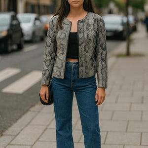 Pode incluir: Uma mulher vestindo uma jaqueta estampada, top preto e jeans azul. A jaqueta tem um design de pele de cobra cinza e preta. Ela também está usando tênis brancos e carregando uma bolsa preta. O fundo mostra uma rua da cidade.