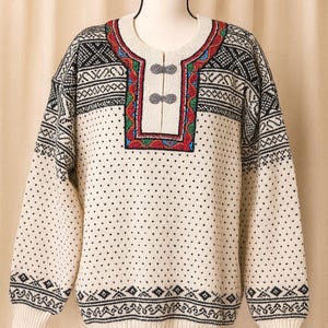 May include: Cream-colored sweater with a detailed geometric pattern in black and white. The neckline features a colorful embroidered design with red, green, and blue accents. Two gray buttons fasten the front.