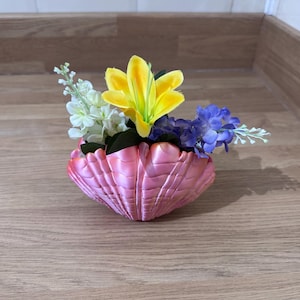 Könnte beinhalten: Ein rosa Muschelkasten mit einer gelben Lilie, weißen Blumen und violetten Blumen.