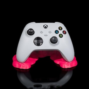May include: A white Xbox controller with black buttons and a pink stand. The controller is resting on the stand, which is shaped like a pair of lips.