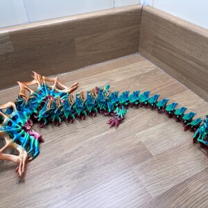 May include: A 3D printed dragon model with a segmented body, featuring a green, blue, and orange color scheme. The dragon is posed in a curled position with its head raised.