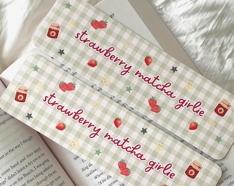 May include: Two rectangular bookmarks featuring a light green and white gingham pattern. The text "strawberry matcha girlie" is printed in a dark pink script font. The design includes small illustrations of red strawberries, jam jars, and stars.