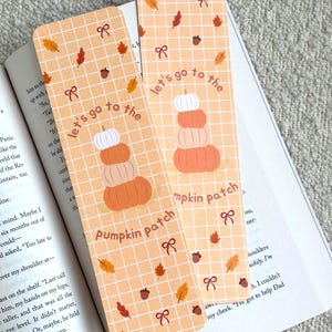 May include: Two rectangular paper bookmarks with a light orange grid pattern, featuring a stack of three pumpkins and the text "let's go to the pumpkin patch" in brown. The design includes small brown acorns, orange leaves, and red bows scattered throughout.