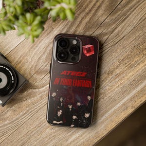 May include: A black smartphone case with the text "ATEEZ IN YOUR FANTASY" in red. The case has a dark background with a group of people and a red crystal. The phone case is on a wooden surface.