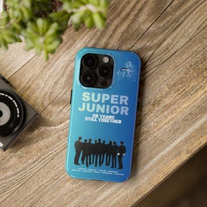 Puede incluir: Funda de teléfono azul con el texto "SUPER JUNIOR 20 YEARS STILL TOGETHER" en blanco. Debajo, una silueta de un grupo de personas. Sobre una superficie de madera.