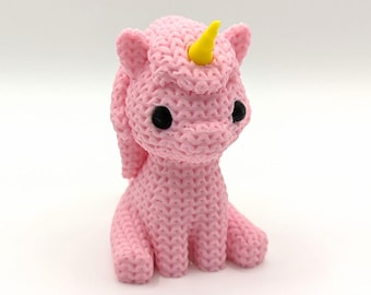 May include: A pink crocheted unicorn figurine with a yellow horn and black eyes. The handmade unicorn has a textured appearance and is sitting upright. The unicorn's mane is styled in a flowing design.