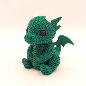 A crochet-textured green dragon toy viewed from the right side, sitting upright with black eyes, white horns, and green wings, giving a three-quarter view of its body.
