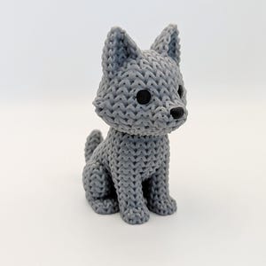 May include: A grey, knitted-style figurine of a wolf or dog. The sculpture has black eyes and a black nose. The animal is sitting upright with its tail curled. The figurine is on a white background.