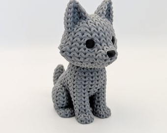 May include: A grey, knitted-style figurine of a wolf or dog. The sculpture has black eyes and a black nose. The animal is sitting upright with its tail curled. The figurine is on a white background.