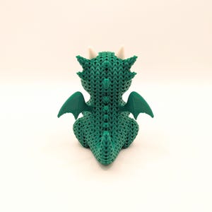 A rear view of a green dragon toy showing its textured back, curved tail, and outstretched green wings. Two white horns are visible on the back of its head.
