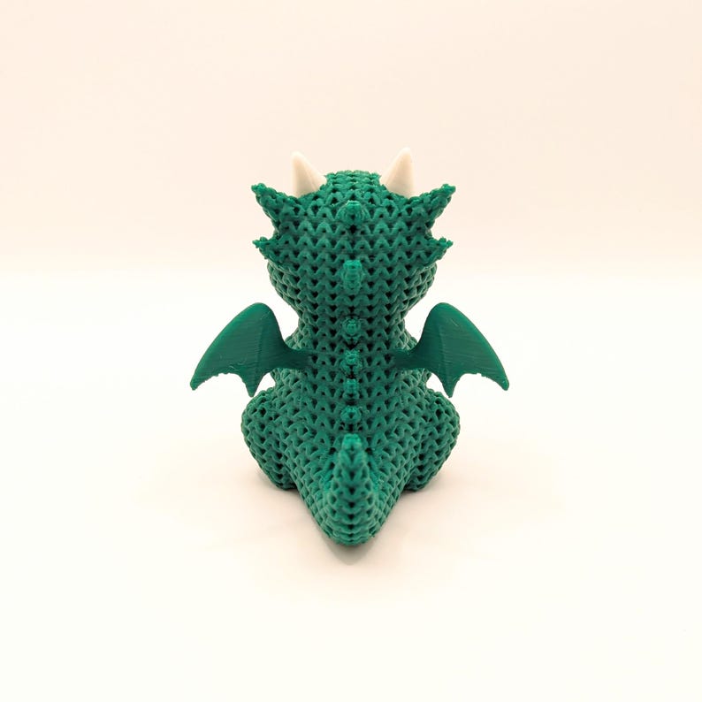 A rear view of a green dragon toy showing its textured back, curved tail, and outstretched green wings. Two white horns are visible on the back of its head.