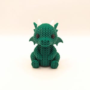 A front-facing view of a cute, green crochet-style dragon toy sitting upright with black eyes, white horns, and small green wings extending from its back.