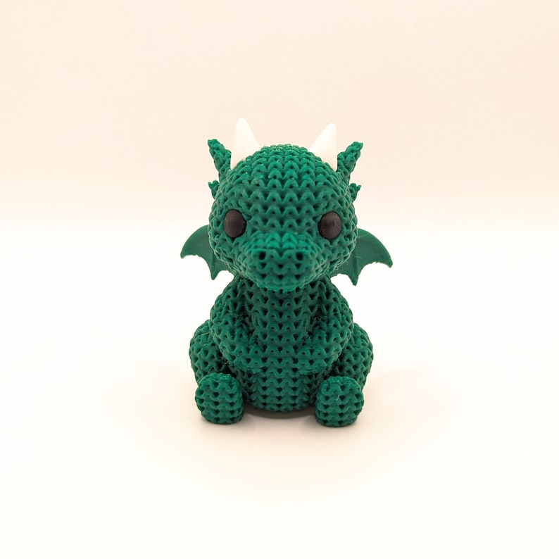 A front-facing view of a cute, green crochet-style dragon toy sitting upright with black eyes, white horns, and small green wings extending from its back.