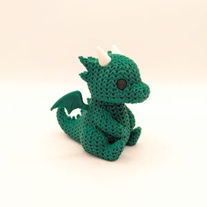 A small, green dragon toy with a crochet-like texture, black eyes, white horns, and green wings. The dragon is sitting upright and facing slightly to the left.