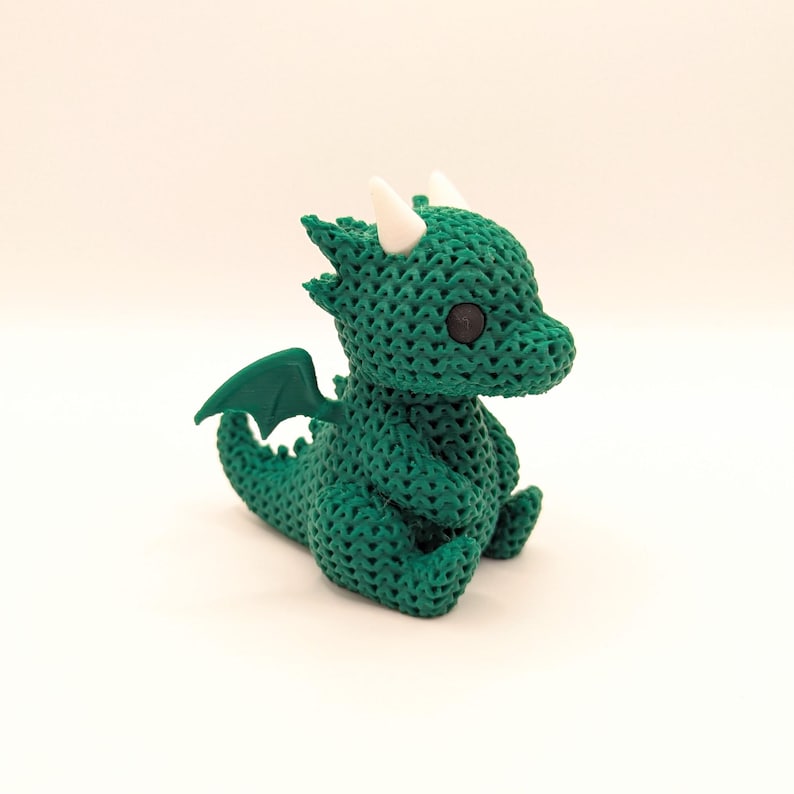 A small, green dragon toy with a crochet-like texture, black eyes, white horns, and green wings. The dragon is sitting upright and facing slightly to the left.