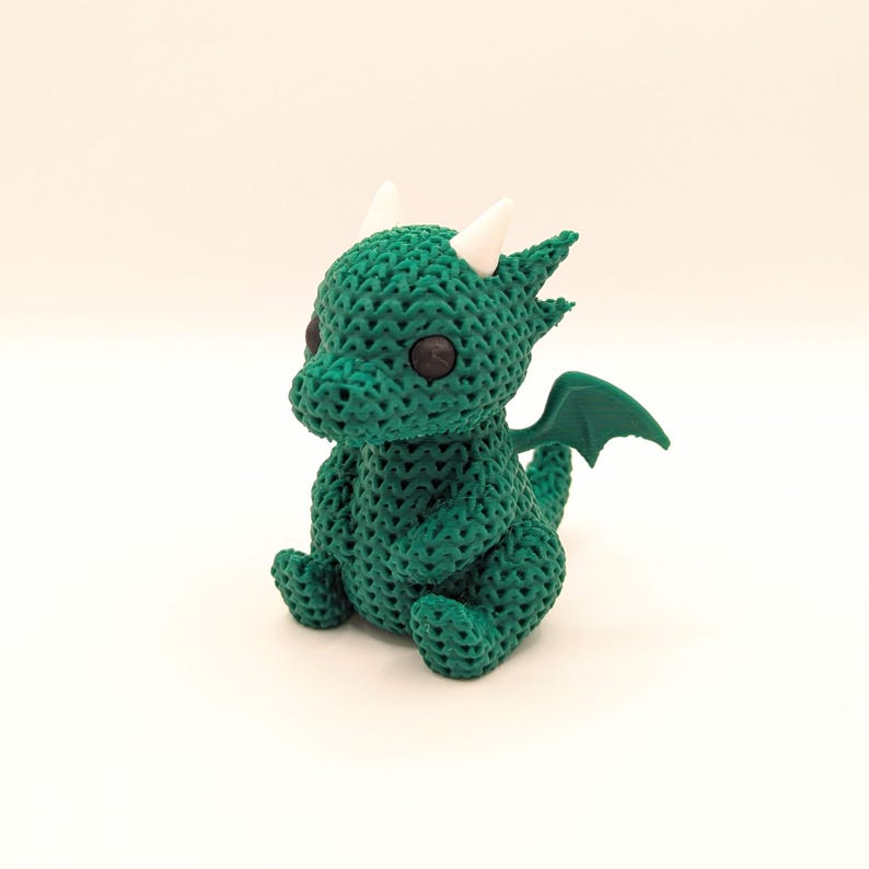 A crochet-textured green dragon toy viewed from the right side, sitting upright with black eyes, white horns, and green wings, giving a three-quarter view of its body.