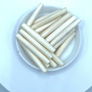 May include: A collection of off-white, cylindrical wooden dowels arranged inside a white, shallow bowl. The dowels are smooth and evenly cut, with a slightly tapered shape. The bowl sits on a white surface, creating a clean, minimalist aesthetic.