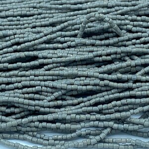 Könnte beinhalten: Nahaufnahme eines Haufens grauer quadratischer Perlen. Die Perlen sind aneinandergereiht und bilden lange Stränge. Die Perlen haben eine einfarbige, matte graue Farbe. Die Perlen sind klein und gleichmäßig groß, ideal für die Schmuckherstellung.