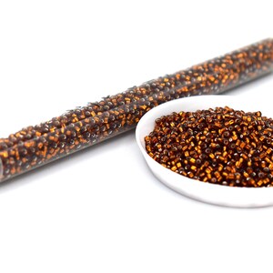 Op de afbeelding: Een doorzichtige plastic buis gevuld met kleine, bruine kralen, naast een witte kom vol met vergelijkbare kralen. De kralen hebben een metallic glans. Ideaal voor het maken van sieraden of knutselen.