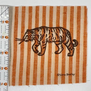 May include: A square piece of fabric featuring vertical orange and white stripes with a dark brown tiger illustration. A measuring tape on the left shows the fabric is approximately 5.5 inches tall. The text Lora Shelley is printed in the corner.