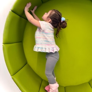 May include: A young child wearing a pastel-colored sweater, gray leggings, and pink boots stands inside a large, lime-green, rounded structure. The child's arms are raised, touching the interior of the structure.