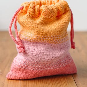 Könnte beinhalten: Eine gehäkelte Kordeltasche mit einem Ombre-Design in Rosa, Orange und Pfirsich. Die Tasche ist aus einem weichen, strukturierten Garn gefertigt und hat eine schlichte, klassische Form.