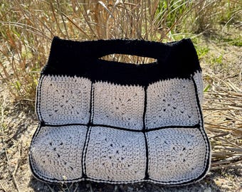 May include: A handmade crochet bag featuring a black handle and trim, with a light gray and black square pattern. The bag is made of soft yarn and has a unique, textured design. The bag is sitting on a sandy surface.