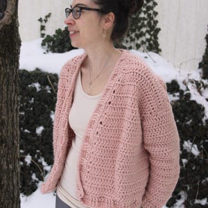 May include: A pink crocheted cardigan with a v-neck and button closure. The cardigan is worn over a white tank top and gray pants.