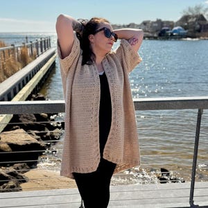 May include: A long, beige crochet cardigan with a loose fit and open front. The cardigan is worn by a person standing on a wooden walkway by the water.