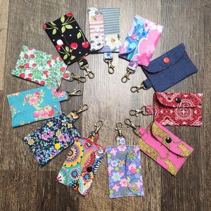 Könnte beinhalten: Eine Sammlung von Kartenhaltern aus Stoff mit Schlüsselanhängern. Die Halter weisen verschiedene Muster auf, darunter Blumen-, Kirsch- und Paisley-Designs. Jeder Halter hat einen Druckknopfverschluss und einen Metallverschluss zum Befestigen an Schlüsseln oder einer Tasche.
