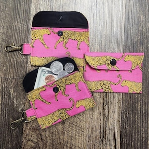 May include: Three pink fabric wallets with a yellow leopard print. One wallet is open, revealing cash and coins. The wallets have black interiors and snap closures. One wallet has a metal clasp attached.