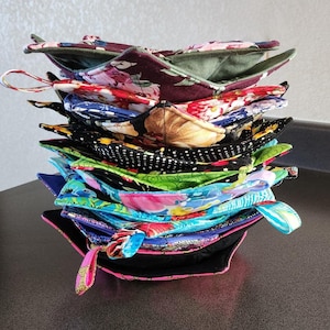 May include: A vertical stack of fabric bowl cozies featuring various patterns including floral prints in burgundy, blue, and pink, black with white polka dots, and bright green designs. Each square-shaped cozy has rounded corners and fabric loops.