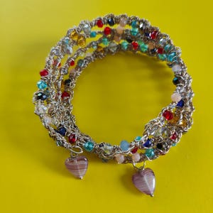 Op de afbeelding: Een armband met meerdere strengen met zilverkleurige draad en kleurrijke kralen in rood, blauw, groen en geel. Twee hartvormige bedels hangen aan de armband. De armband wordt op een gele achtergrond gepresenteerd.