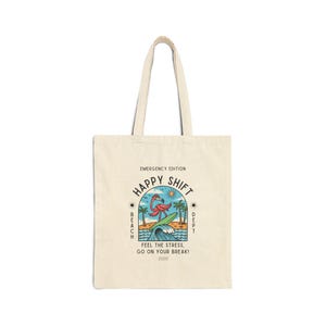 May include: Cream-colored tote bag with a tropical design. The design features a flamingo surfing a wave, palm trees, and the words "HAPPY SHIFT" and "FEEL THE STRESS, GO ON YOUR BREAK!" in a circular layout. The bag has two handles.