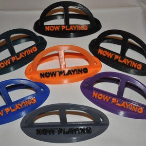 May include: Six different colored plastic holders with the words "Now Playing" printed in orange on each. The holders are shaped like a half-circle with a small opening in the center.