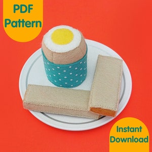May include: A felt craft pattern for a boiled egg and two slices of toast on a white plate. The egg is teal blue with white polka dots and a yellow yolk. The toast is beige. The pattern is available for instant download. The text "PDF Pattern" is in a yellow circle and the text "Instant Download" is in a yellow rectangle.