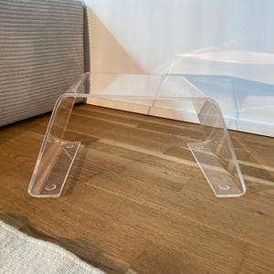 Puede incluir: Una mesa de acrílico transparente con un diseño curvo. La mesa está hecha de una sola pieza de acrílico y tiene un diseño simple y moderno.