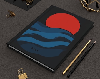 May include: Black journal with a red sun and blue wave design. The journal has the text "kira j." on the cover. Gold and black pencils and gold binder clips are also visible.